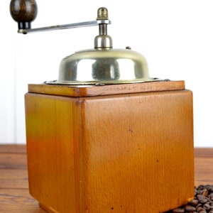 K&ouml;nnte beinhalten: Eine antike Kaffeem&uuml;hle aus Holz mit silbernem Oberteil und Handkurbel. Die M&uuml;hle steht auf einer Holzoberfl&auml;che, um die herum Kaffeebohnen verstreut liegen.