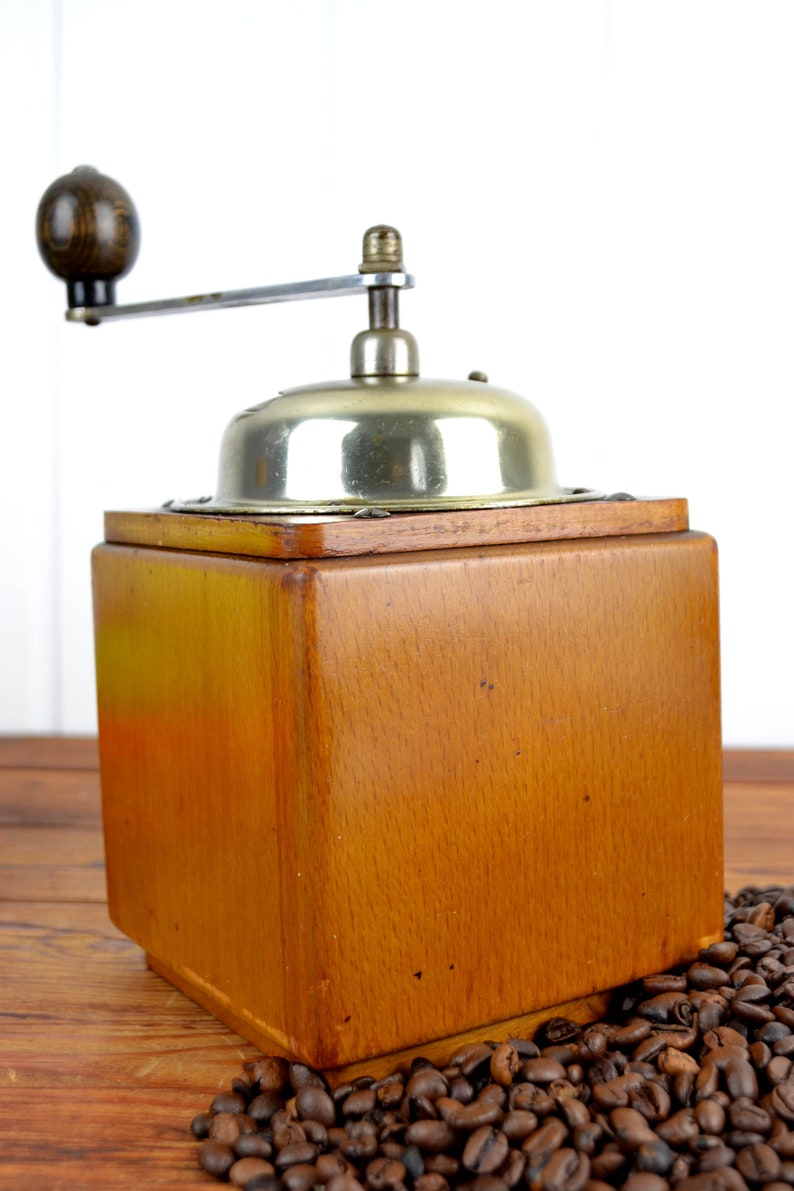 K&ouml;nnte beinhalten: Eine antike Kaffeem&uuml;hle aus Holz mit silbernem Oberteil und Handkurbel. Die M&uuml;hle steht auf einer Holzoberfl&auml;che, um die herum Kaffeebohnen verstreut liegen.