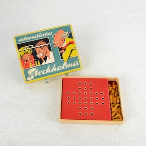 May include: A vintage game box with a red and blue illustration of people playing a game. The box is labeled "Schwedisches Steckhalma". The game includes a red game board with holes and a pile of small wooden pieces.