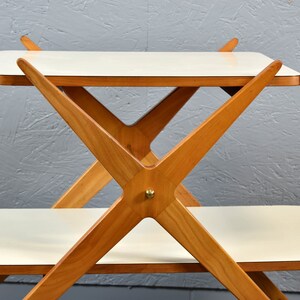 May include: A two-tiered white serving table with a wooden frame. The frame is made of light brown wood and has a criss-cross design. The table has a white top and a white bottom shelf.