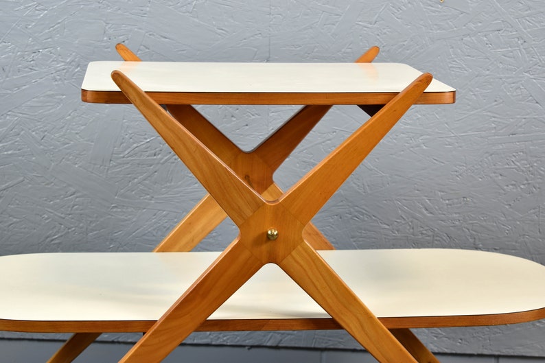 May include: A two-tiered white serving table with a wooden frame. The frame is made of light brown wood and has a criss-cross design. The table has a white top and a white bottom shelf.