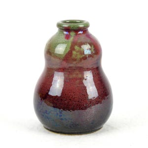 Puede incluir: Un jarrón de cerámica hecho a mano con un cuerpo bulboso y un cuello estrecho. El jarrón presenta un acabado brillante con una mezcla de colores, incluyendo tonos rojo oscuro, verde y azul. El jarrón mide aproximadamente 15 cm de alto.