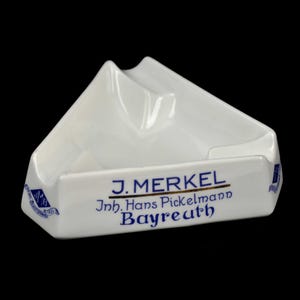 Könnte beinhalten: Weißer Keramik-Aschenbecher mit dreieckigem Design. Der Aschenbecher trägt den Text "J. Merkel, Inh. Hans Pickelmann, Bayreuth" in blauen und goldenen Buchstaben. Der Aschenbecher hat eine glatte, glänzende Oberfläche.