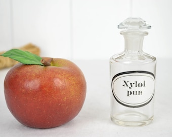 May include: A red apple with a green leaf next to a clear glass bottle with a stopper. The bottle has a label that reads "Xylol pur". The background is white.