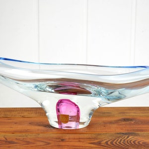 May include: A clear glass bowl with a blue rim and a pink glass base. The bowl is shaped like a wave and has a unique, organic design.