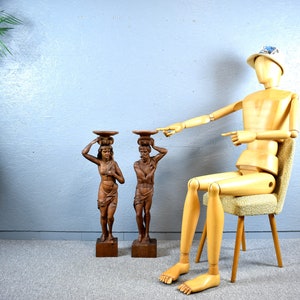 Puede incluir: Dos esculturas de madera de personas que llevan cestas en la cabeza. Las esculturas est&aacute;n sobre bases de madera. Un maniqu&iacute; de madera est&aacute; sentado en una silla en el fondo.