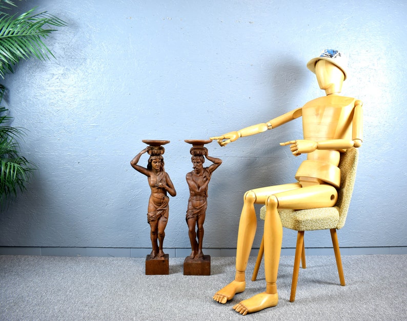 Puede incluir: Dos esculturas de madera de personas que llevan cestas en la cabeza. Las esculturas est&aacute;n sobre bases de madera. Un maniqu&iacute; de madera est&aacute; sentado en una silla en el fondo.