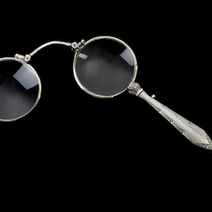Könnte beinhalten: Antike Brille aus Silber mit zwei runden Gläsern und einem dekorativen Griff. Der Griff weist aufwendige Gravuren und eine kleine Schlaufe auf. Die Gläser sind klar, und der Rahmen ist silberfarben. Der Hintergrund ist schwarz.