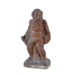 Könnte beinhalten: Verwitterte, braune Statue eines bärtigen Mannes, möglicherweise eine mythologische Figur, die auf einem rechteckigen Sockel steht. Die Statue hat eine raue Textur und ist mit einem langen Bart und drapiertem Stoff dargestellt. Die Figur hält einen Gegenstand in der linken Hand.