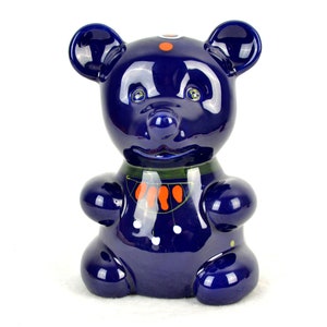 Può includere: Un salvadanaio in ceramica blu a forma di orso con dettagli dipinti in arancione, verde e bianco.