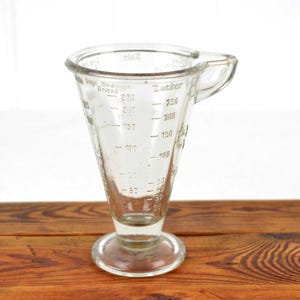 May include: Clear glass measuring jug with a flared cone shape, a handle, and a round base. The jug has measurement markings in millilitres and ounces. The word "Zucker" is printed on the side. It sits on a wooden surface.