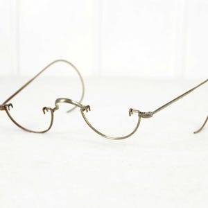 May include: Antique wire-rimmed eyeglasses with a delicate, minimalist design. The glasses feature a curved bridge and temples, and are made of a thin, silver-toned metal. The lenses are missing.