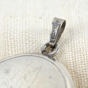 May include: Close-up of a silver pendant with a loop marked "Alpaca". The pendant has a circular design with visible scratches. The background is a textured, off-white fabric.