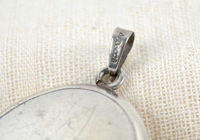 May include: Close-up of a silver pendant with a loop marked "Alpaca". The pendant has a circular design with visible scratches. The background is a textured, off-white fabric.
