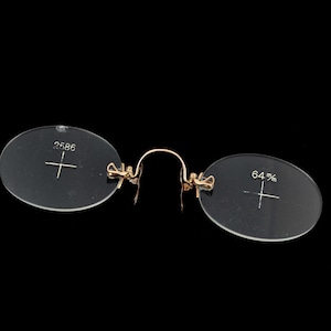 Puede incluir: Gafas antiguas de pinza con lentes ovaladas transparentes y un puente dorado. La lente izquierda tiene el número "2586" y una retícula, mientras que la lente derecha tiene "64%" y una retícula. Las gafas están sobre un fondo negro.