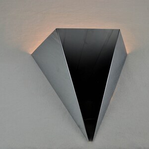 May include: A silver triangular wall sconce with a black interior. The sconce is mounted on a white wall.