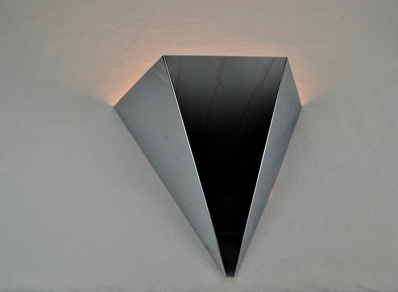 May include: A silver triangular wall sconce with a black interior. The sconce is mounted on a white wall.