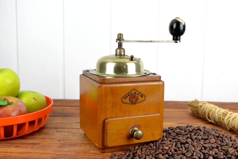 K&ouml;nnte beinhalten: Eine antike Kaffeem&uuml;hle aus Holz mit Messingdeckel und schwarzem Griff. Die M&uuml;hle hat eine Schublade zum Auffangen des Kaffeesatzes und ein Etikett mit der Aufschrift "Dresden".