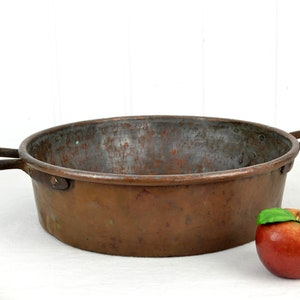 Puede incluir: Una olla de cobre grande y redonda con dos asas de hierro. La olla tiene una pátina desgastada y envejecida y está sobre una superficie blanca. Una manzana roja con una hoja verde está en primer plano.