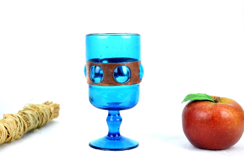 May include: A blue glass goblet with a copper band and a round base. The goblet is filled with a clear liquid.