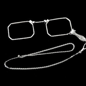 Può includere: Un paio di occhiali argentati con lenti rettangolari, collegati da un ponte e un'impugnatura decorativa. Una delicata catena è attaccata, probabilmente per essere indossata al collo. Sfondo nero.