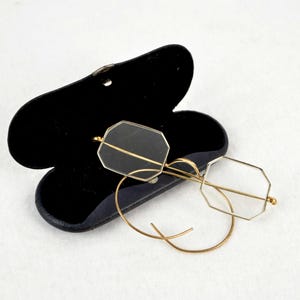 以下が含まれることがあります： 金色の縁取りが施された八角形の眼鏡が、黒い開いた眼鏡ケースの上に置かれています。眼鏡は繊細なワイヤーフレームのデザインです。ケースは開いており、柔らかく暗い内側が見えます。背景は無地の白い表面です。