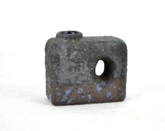 May include: A small, dark gray ceramic vase with a rough texture and blue speckles. The vase has a round opening at the top and a small oval opening on the side.