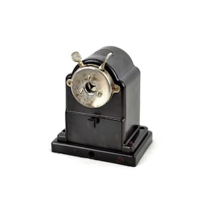 Pode incluir: Um apontador de lápis vintage preto com uma lâmina de metal e uma gaveta para aparas. O apontador tem uma manivela no lado para girar a lâmina.
