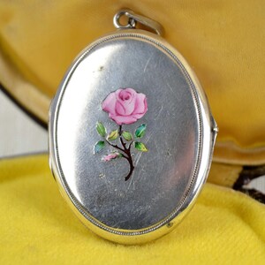 May include: Oval silver locket with a pink rose painted on the front. The rose has green leaves and a brown stem. The locket is open, revealing a yellow interior.