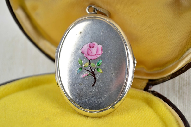 May include: Oval silver locket with a pink rose painted on the front. The rose has green leaves and a brown stem. The locket is open, revealing a yellow interior.