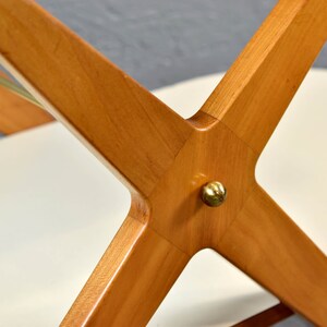 May include: Close-up of a wooden table base with a brass accent. The base is made of four pieces of wood that intersect in the center, forming an X shape. The wood is a light brown color and has a smooth finish.