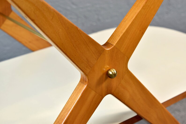 May include: Close-up of a wooden table base with a brass accent. The base is made of four pieces of wood that intersect in the center, forming an X shape. The wood is a light brown color and has a smooth finish.