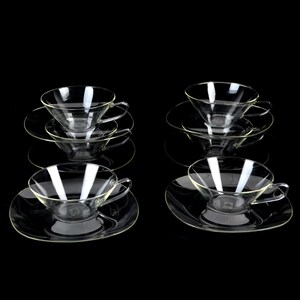May include: Six clear glass teacups and saucers are arranged on a black surface. The teacups have a rounded bowl shape with a handle. The saucers are round and flat. The set is ideal for serving tea or coffee.