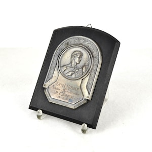 Könnte beinhalten: Eine silberne Plakette mit einem Relief eines Mannes, der einen Lorbeerzweig hält. Die Plakette ist auf einem schwarzen Holzsockel montiert und trägt die Inschrift "DEM SIEGER" und "Wien - 1927 - Wiener B.C. Sport" in deutscher Sprache.