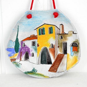 Op de afbeelding: Een keramische wandhanger met een geschilderd tafereel van een klein Italiaans dorp met kleurrijke gebouwen, een blauwe zee en een groene cipres. De hanger heeft een rood touwtje om op te hangen.