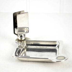 Pode incluir: Um cinzeiro prateado com um porta-cigarros embutido. O cinzeiro tem uma base retangular com cantos arredondados e um pequeno detalhe circular. O porta-cigarros é um design vertical de dois lados.