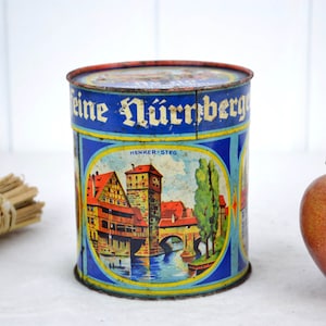 May include: A vintage blue and yellow tin can with a picture of a bridge and buildings. The text on the can reads "Feine Nürnberger" and "Henker-Steg".