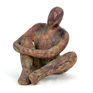 Könnte beinhalten: Eine Keramikskulptur einer Person, die mit gekreuzten Beinen und um die Knie geschlungenen Armen sitzt. Die Skulptur ist aus rotbraunem Ton mit grünen und schwarzen Akzenten gefertigt.