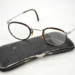 May include: A pair of round, tortoiseshell glasses with silver metal frames and clear lenses. The glasses are resting on a black leather case.