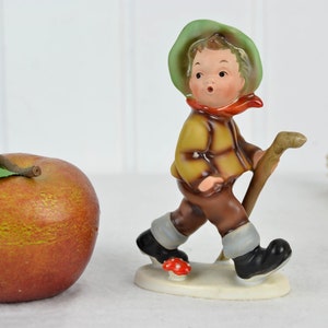 Puede incluir: Una figurita de porcelana de un niño con un sombrero verde, una chaqueta marrón y pantalones marrones. Lleva un palo y camina con un hongo rojo a sus pies.