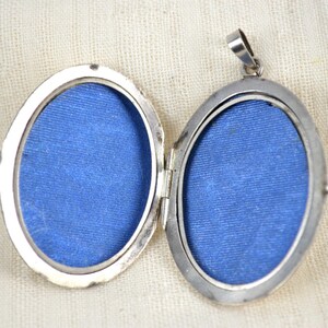 May include: Open silver oval locket with a hinged design. The locket is open, revealing two blue fabric-lined compartments. The locket has a small loop at the top for a chain. The background is a cream-colored fabric.