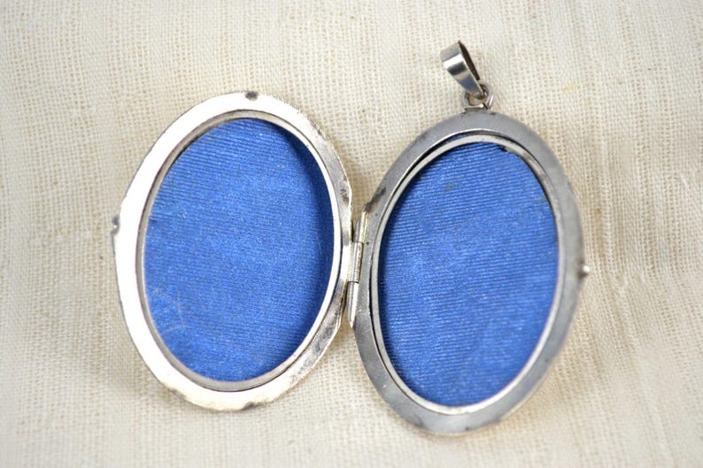May include: Open silver oval locket with a hinged design. The locket is open, revealing two blue fabric-lined compartments. The locket has a small loop at the top for a chain. The background is a cream-colored fabric.