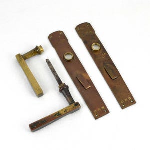 May include: Antique door hardware set, including two long, rectangular bronze plates with circular accents, a brass door handle, and a smaller handle with a dark finish. The pieces are arranged on a white background.