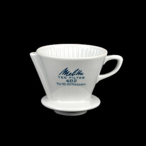 Puede incluir: Filtro de té Melitta 402 de cerámica blanca, con asa y base pequeña. El filtro tiene forma cónica con estrías verticales en el interior. El texto "Melitta TEE FILTER 402 für 10-20 Tassen" está impreso en azul.