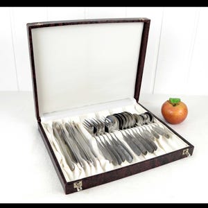 May include: A vintage silverware set in a brown leatherette case. The set includes forks, knives, and spoons, neatly arranged on a white fabric lining. An apple with a green leaf sits beside the open case.