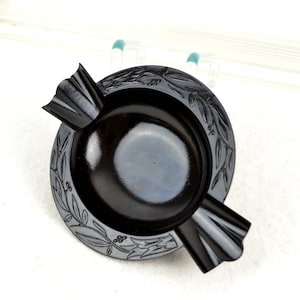 May include: Black ashtray with a decorative design of leaves and berries around the edge. The ashtray has two wings on either side.