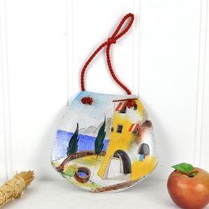 May include: A ceramic wall hanging with a red rope for hanging. The wall hanging depicts a yellow building with red and white striped awnings, green trees, and a blue sea.