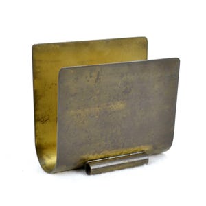 Könnte beinhalten: Ein Vintage-Serviettenhalter aus Messing mit geschwungenem Design. Der Halter hat eine rechteckige Form mit geschwungener Basis und zylindrischer Stütze. Das Messing hat ein angelaufenes, gealtertes Aussehen, was darauf hindeutet, dass es sich um einen antiken oder Vintage-Artikel handelt.