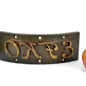 Könnte beinhalten: Ein gebogenes, dunkelbraunes Metallschild mit goldenen Buchstaben, auf dem "OKF3" steht. Das Schild hat sechs gleichmäßig verteilte Löcher entlang der oberen und unteren Kante.