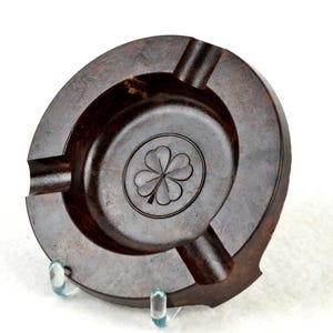 May include: A dark brown ashtray with a circular design. The ashtray features three cigarette rests and a central recessed area with a carved four-leaf clover. The ashtray is made of a dark, possibly wooden material.